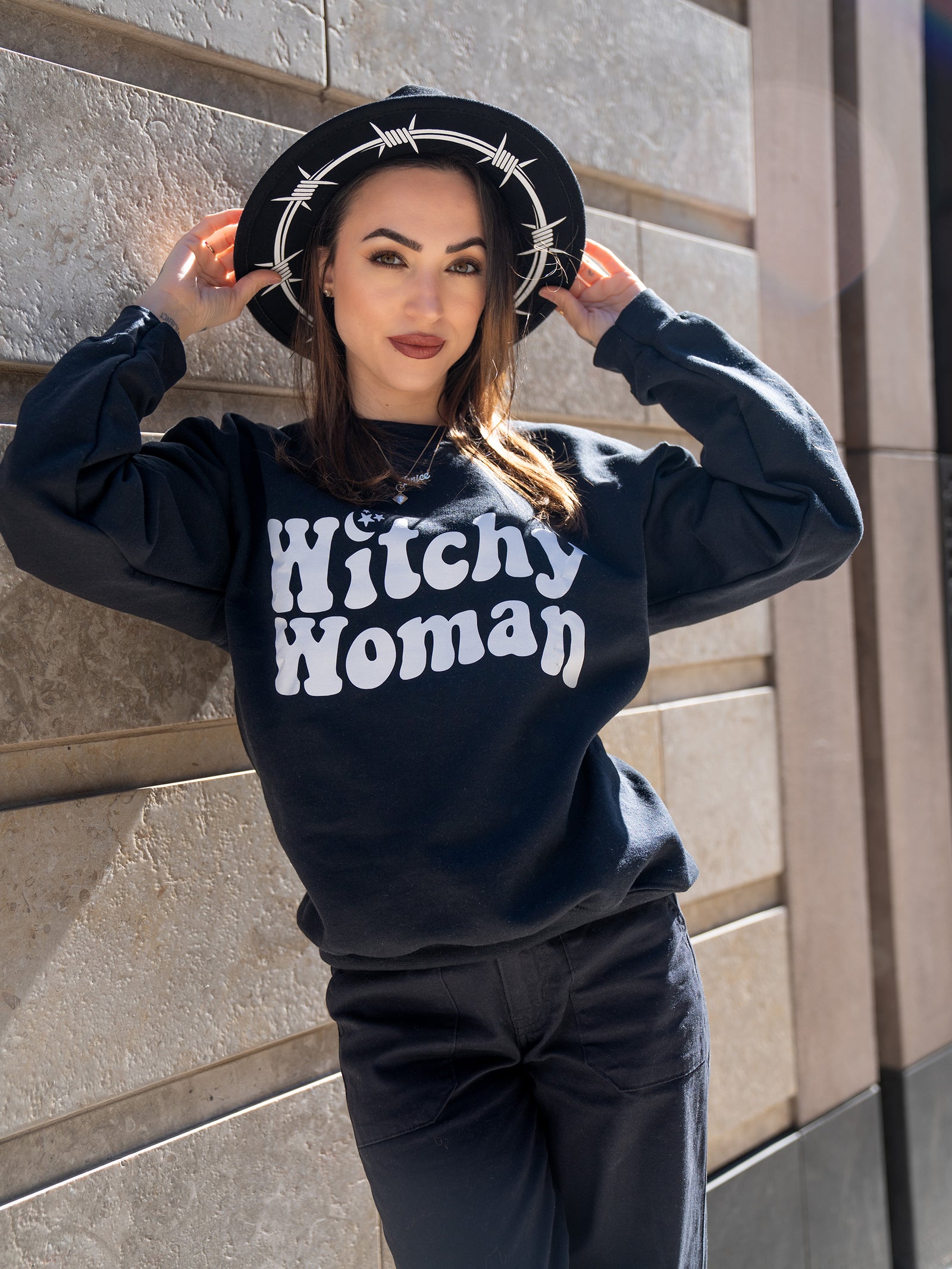 Janice Danger wearing a black wide brim felt hat with Barbed Wire white graphics and the Witchy Woman black crewneck sweatshirt. 