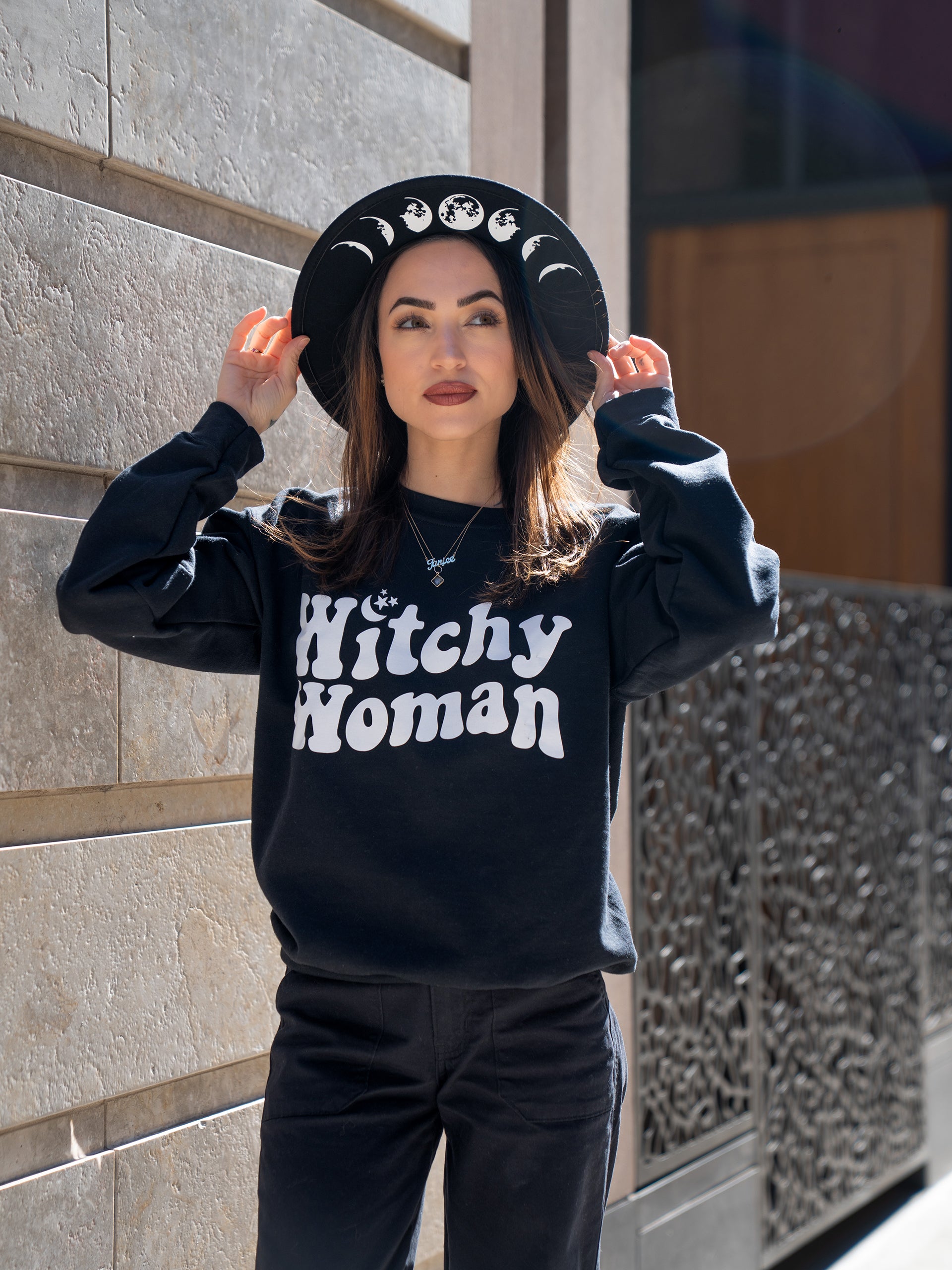 Janice Danger wearing a wide brim felt hat with Moon Phase graphics and the Witchy Woman black crewneck sweatshirt. 