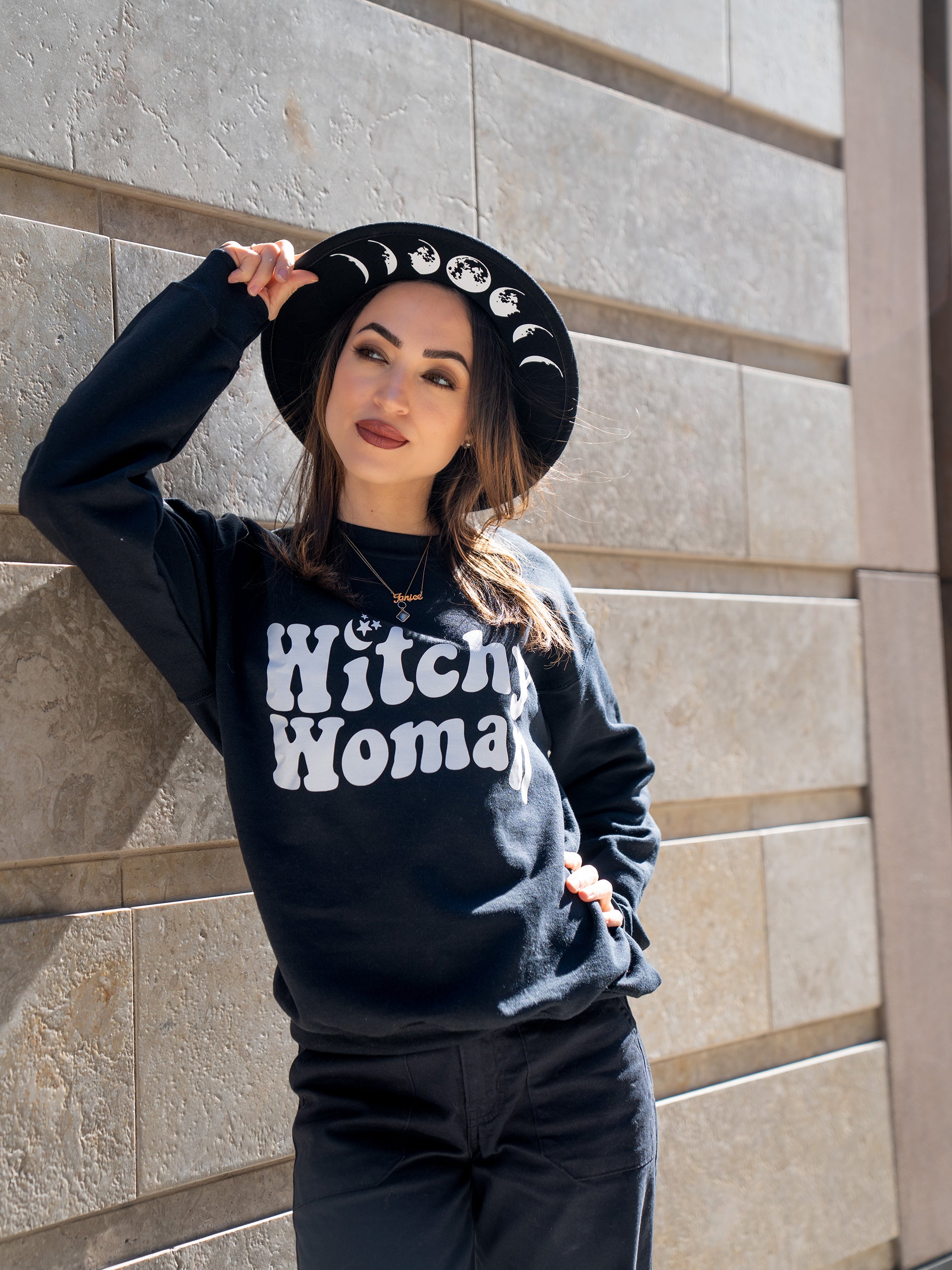 Janice Danger wearing a wide brim felt hat with Moon Phase graphics and the Witchy Woman black crewneck sweatshirt. Photographed against a stone wall in NYC.