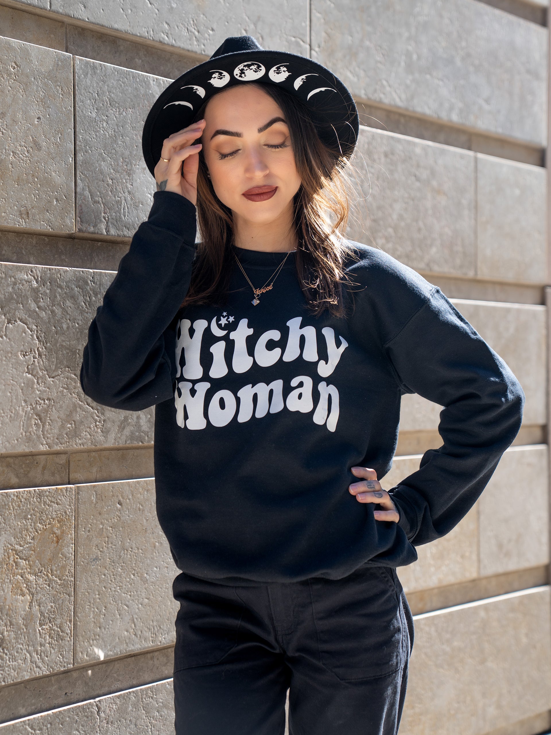 Janice Danger wearing a wide brim felt hat with Moon Phase graphics and the Witchy Woman black crewneck sweatshirt. 