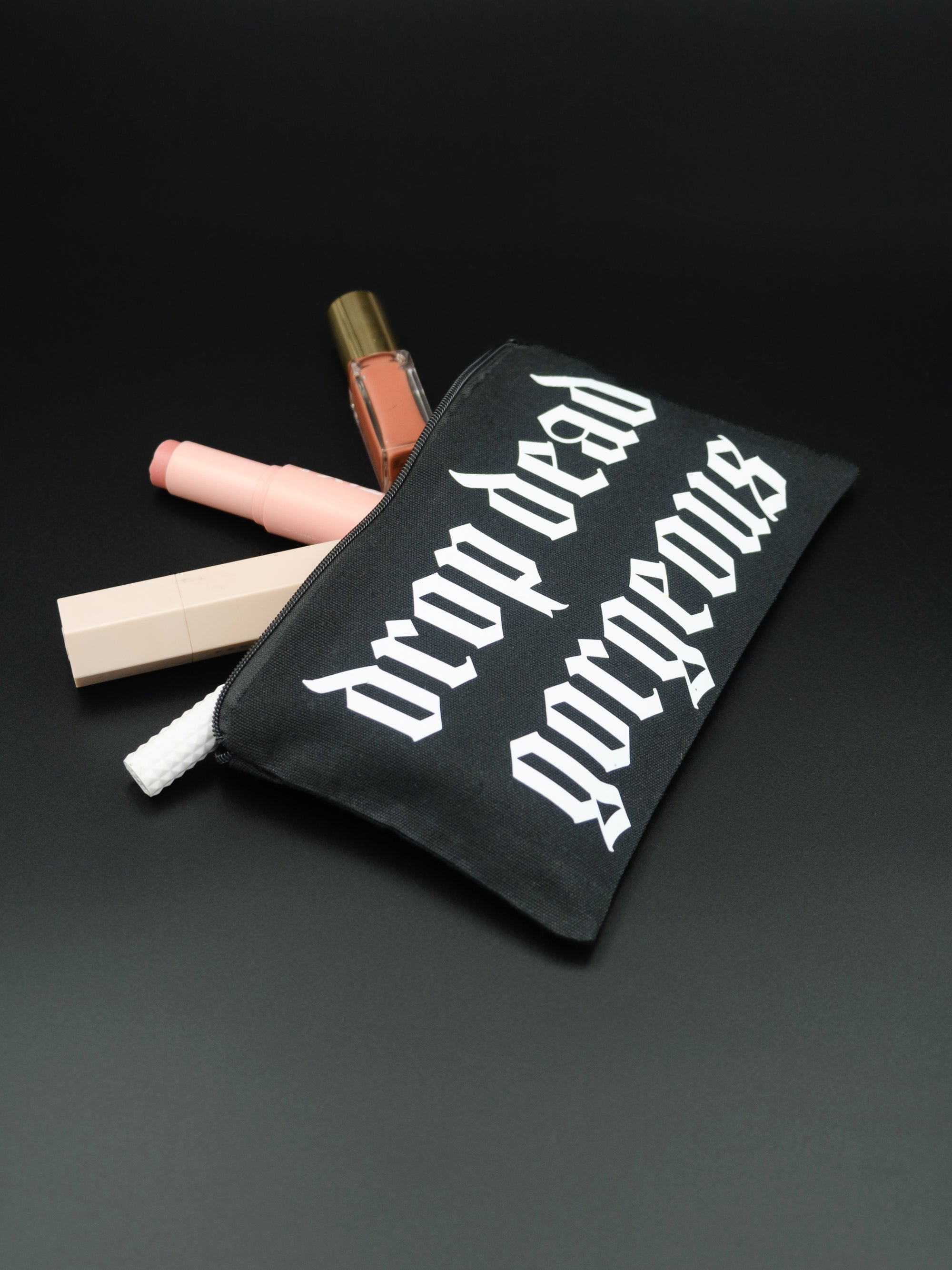 Angled shot of a black cosmetic bag with cosmetics on a black background. zip pouch reads Drop Dead Gorgeous in white Old English style text. 