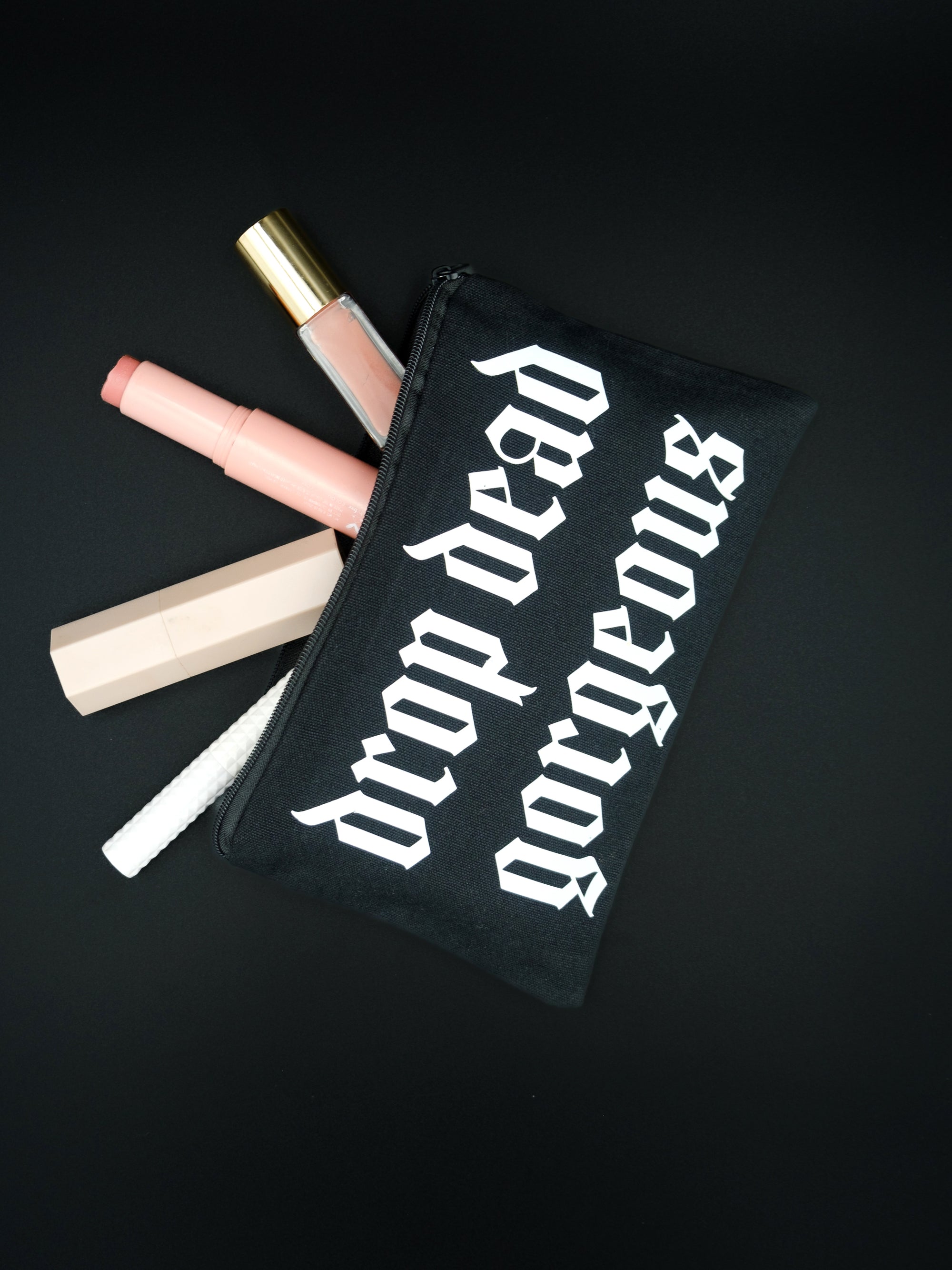 Black cosmetic bag with cosmetics on a black background. zip pouch reads Drop Dead Gorgeous in white Old English style text. 