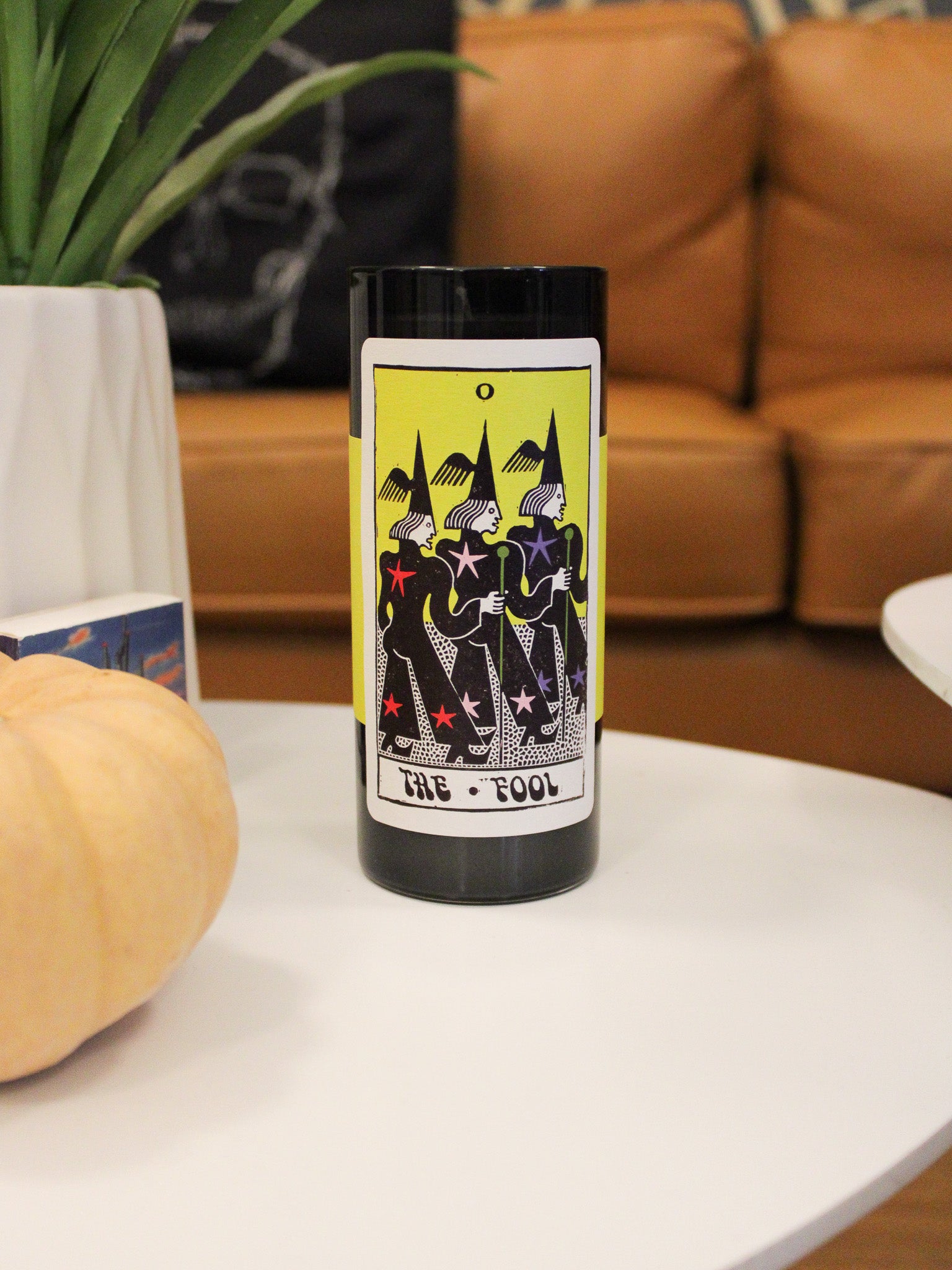 black glass tall votive candle with tarot card style label reading "The fool". Features an illustration of three people. 