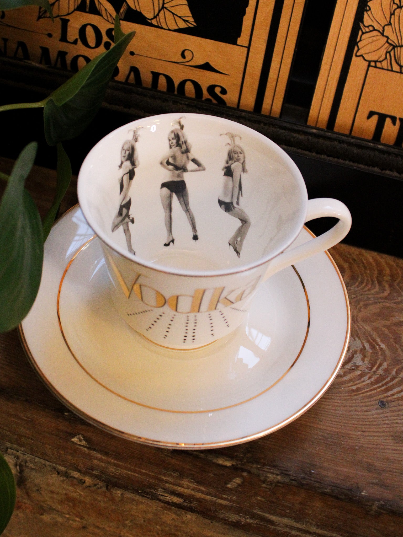 White china teacup with gold detailing reads "vodka" with grayscale dancing women on the inner cup and gold trim. 