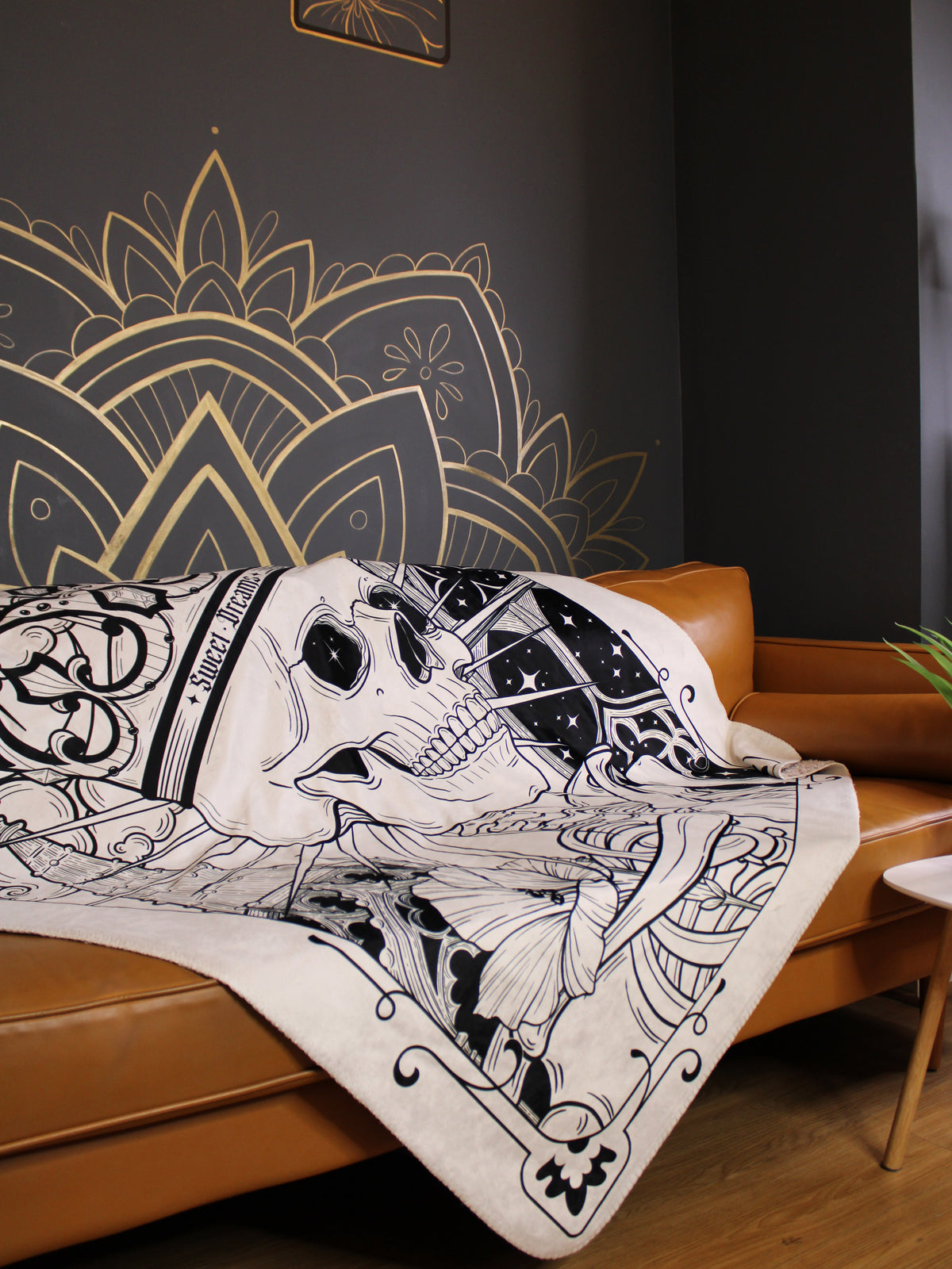 White soft blanket with skull and architectural designs made by Janice Danger draped over a caramel colored couch against a gray wall at Grit N Glory in NYC