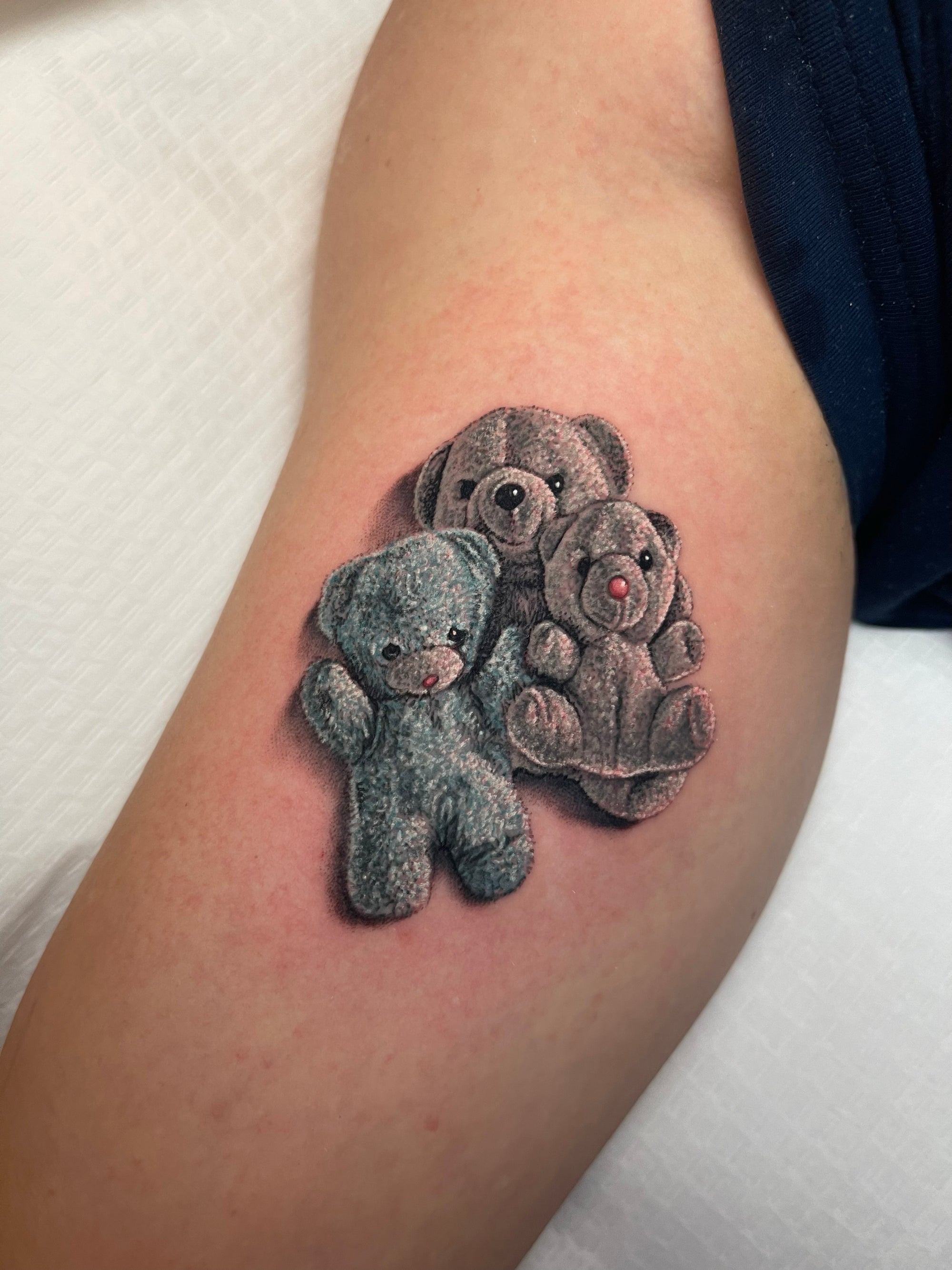 Color realism stuffed animal upper arm tattoo by Megan Massacre at Grit n Glory in New York City. This realistic small tattoo features one blue teddy bear with two grey teddy bears.