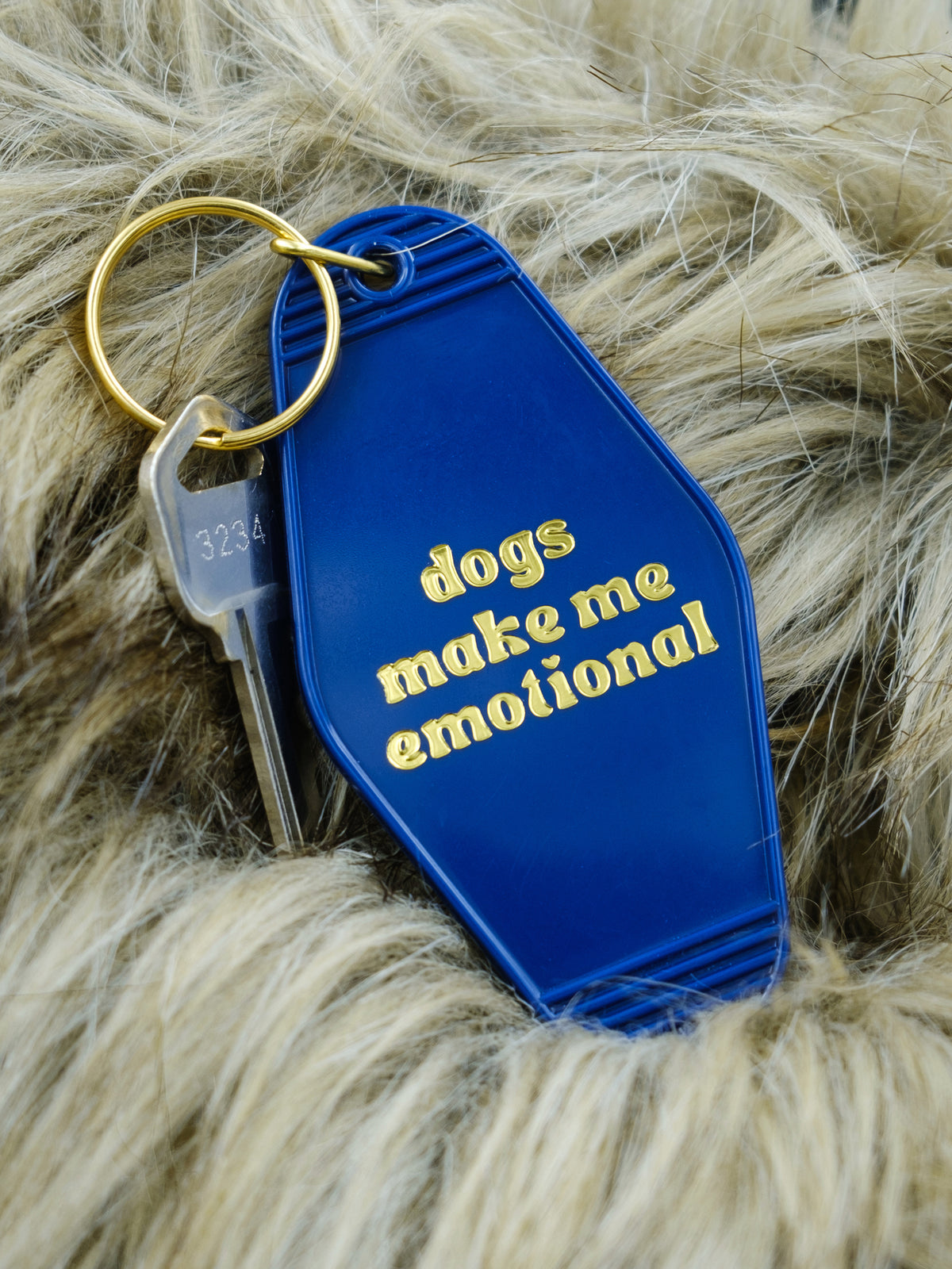 blue motel style keychain reads &quot;dogs make me emotional&quot; in gold, pictured against a faux fur background