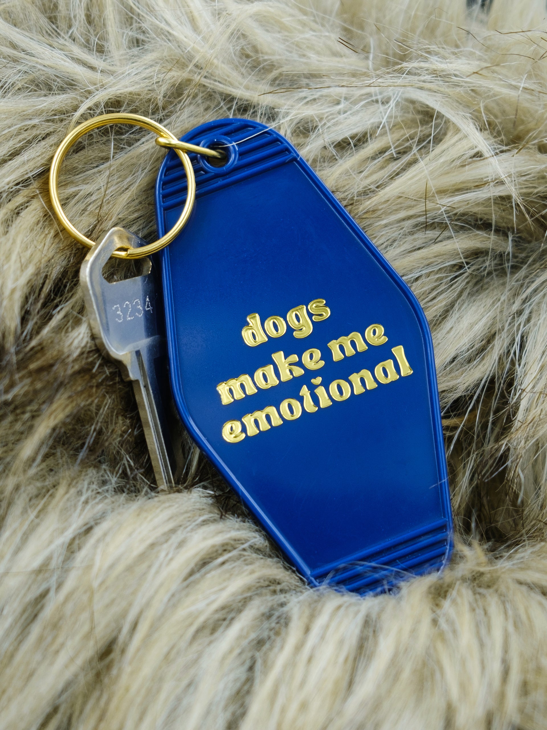 blue motel style keychain reads "dogs make me emotional" in gold, pictured against a faux fur background