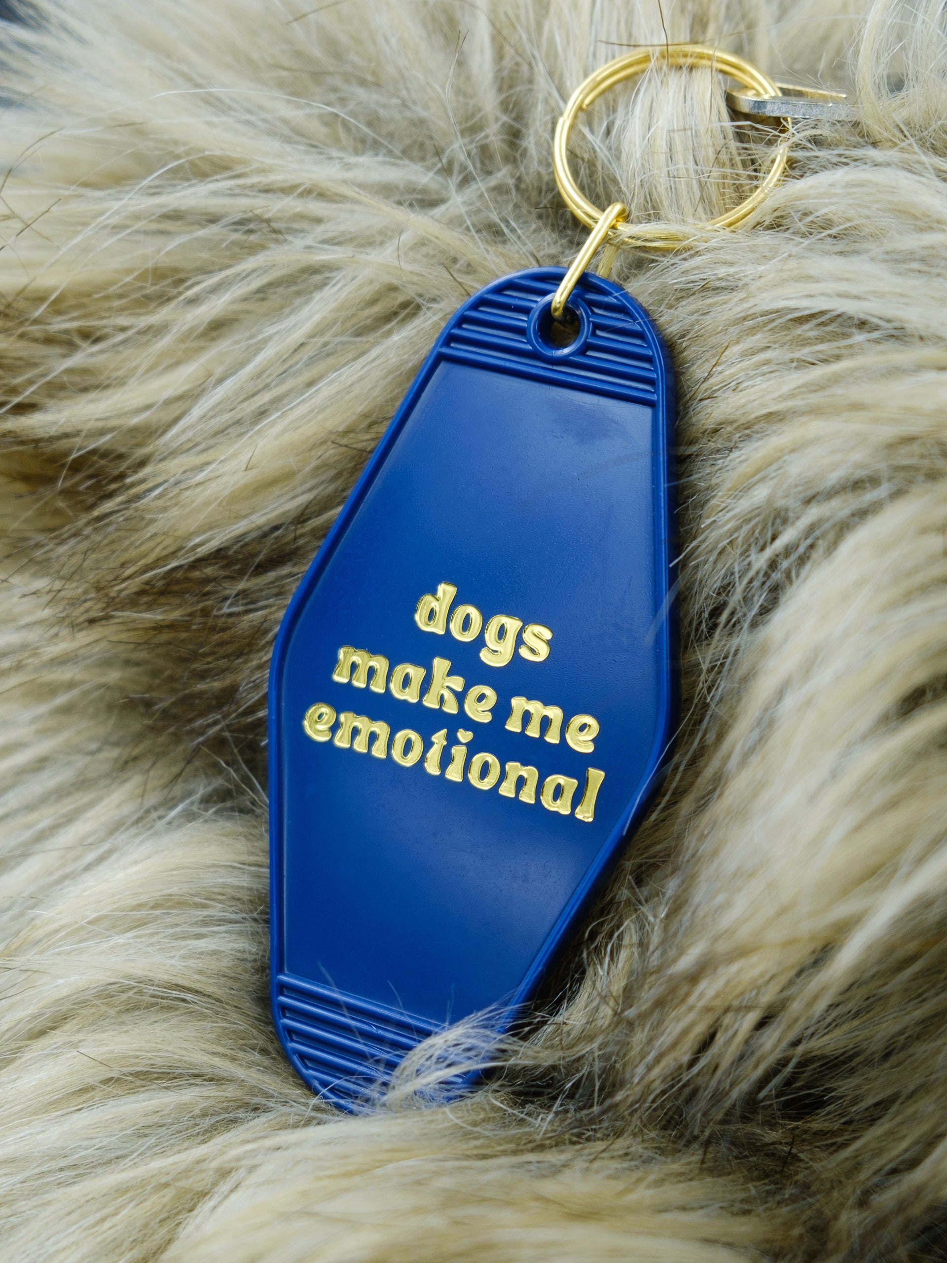 blue motel style keychain reads "dogs make me emotional" in gold, pictured against a faux fur background