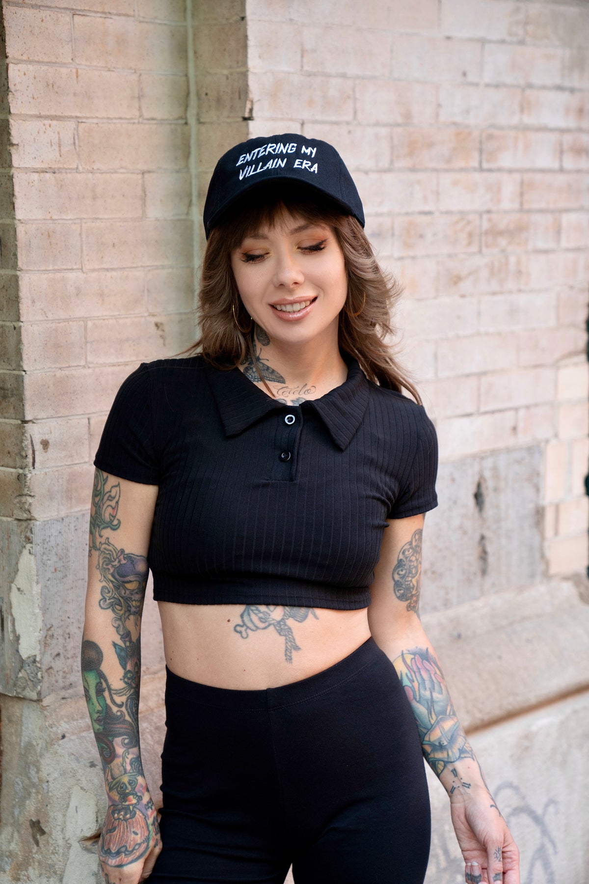 Megan Massacre wearing the Villain Era black and white dad hat with a crop top against a brick wall.