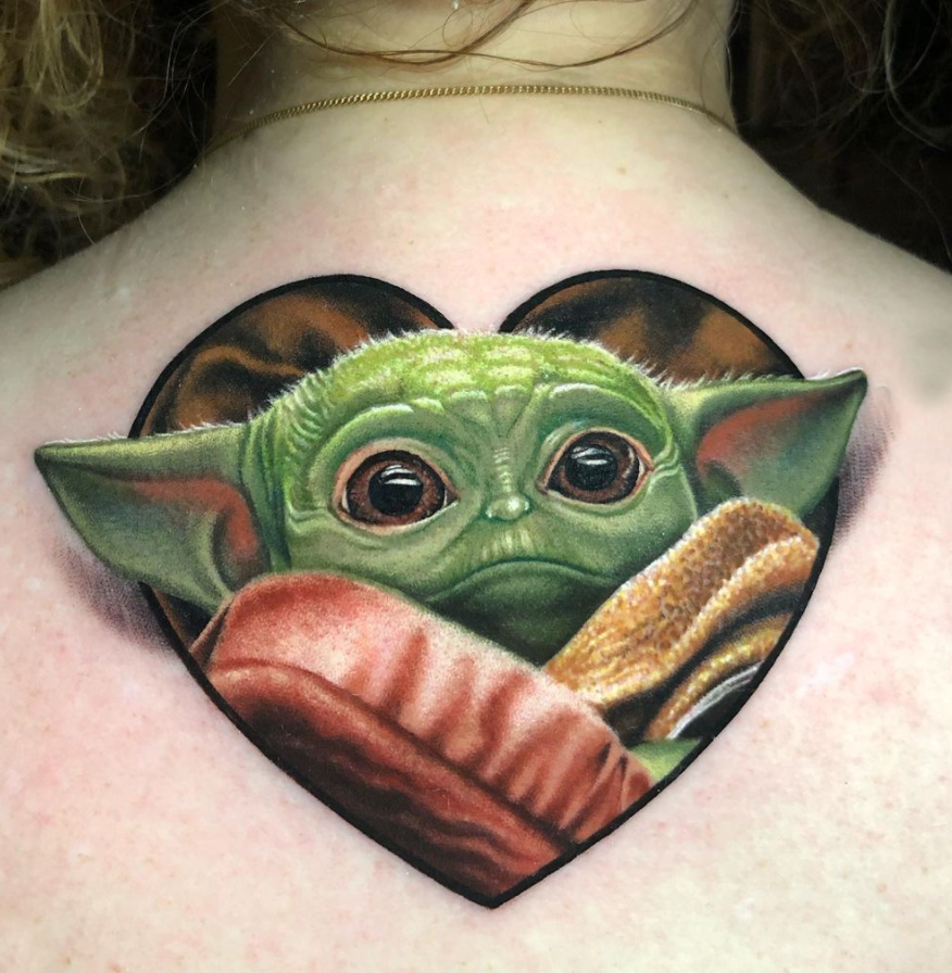 Color realism Baby Yoda, Grogu Tattoo on the upper back by Megan Massacre. This cute realistic tattoo features heart shaped frame with Grogu's ears sticking out for a 3d effect. 