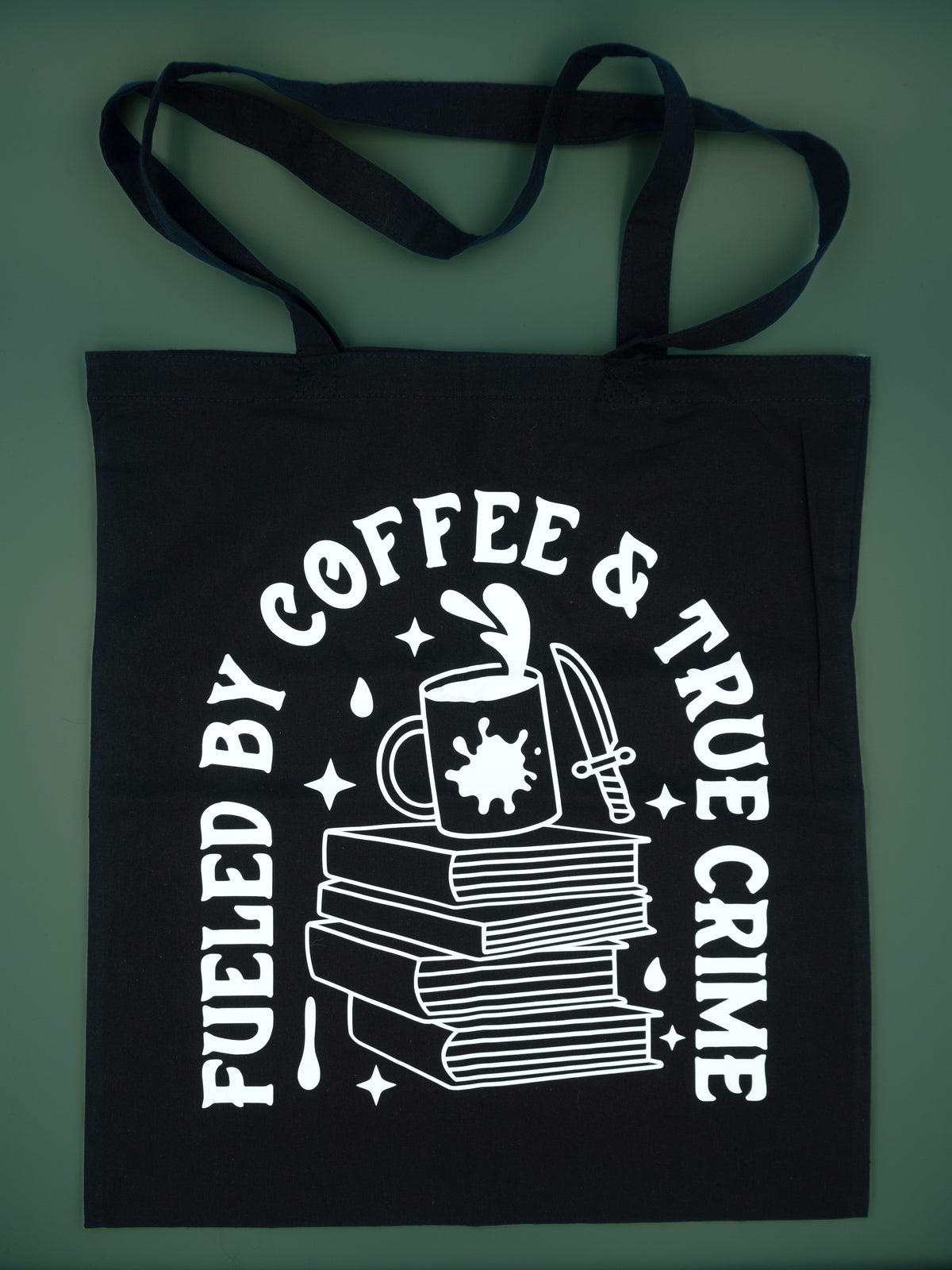 Black canvas totebag on a green background, tote bag reads Fueled By Coffee &amp; True Crime