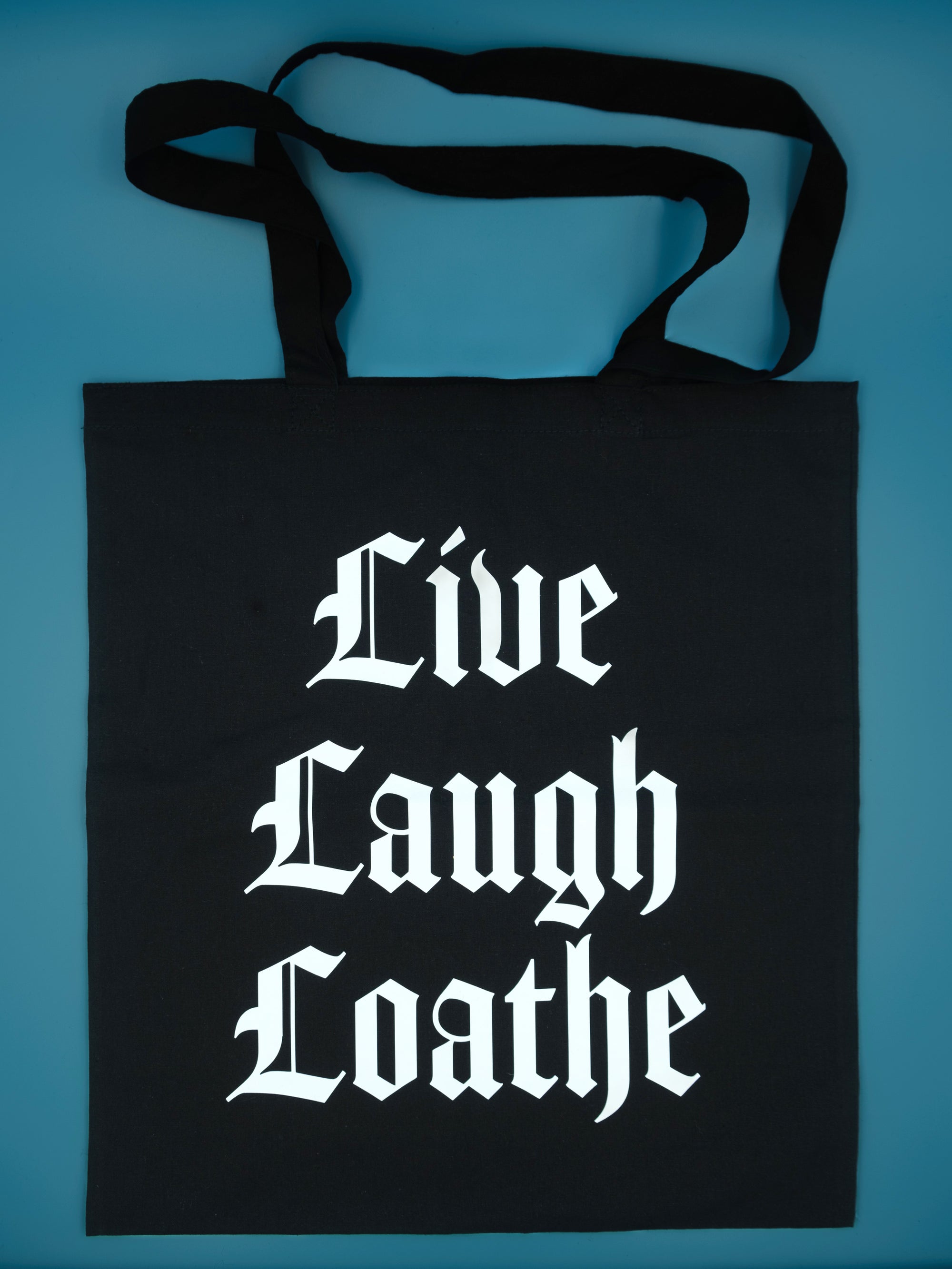 Black canvas totebag on a blue background, tote bag reads  Live Laugh Loathe in white 