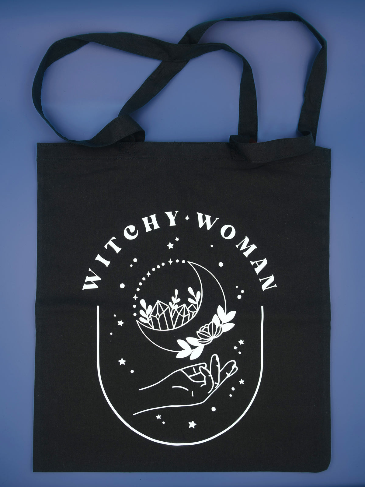 Black canvas totebag on a blue background, tote bag reads Witchy Woman with a crescent moon 