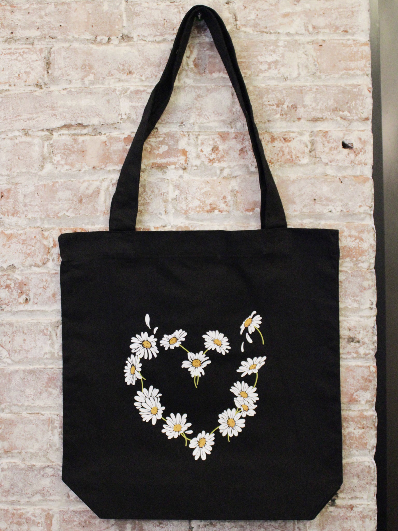 black cotton tote bag with daisy heart shaped graphic photographed against a brick wall. 