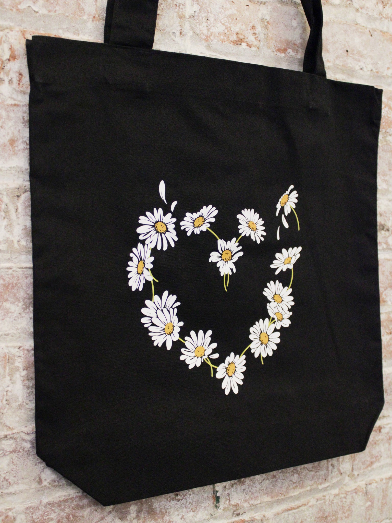 black cotton tote bag with daisy heart shaped graphic photographed against a brick wall. 