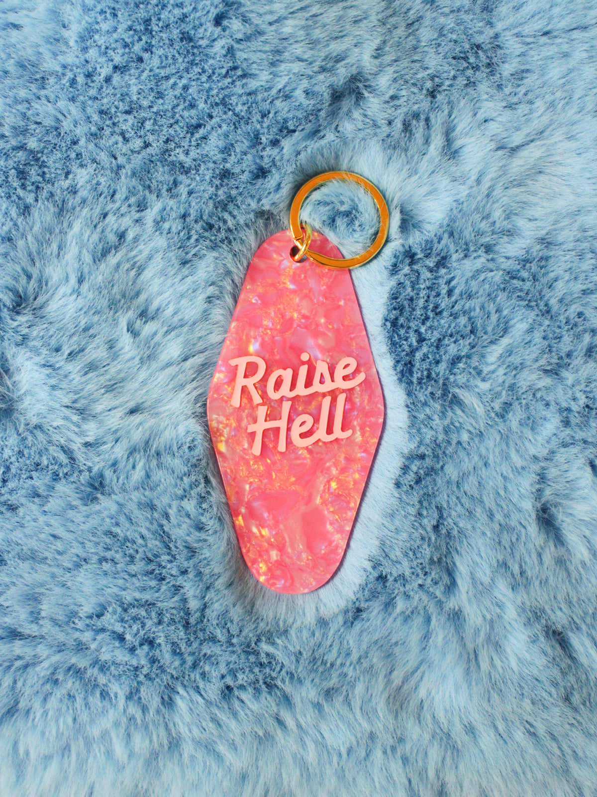 pink motel style keychain reads &quot;raise hell&quot; photographed on a fuzzy blue background