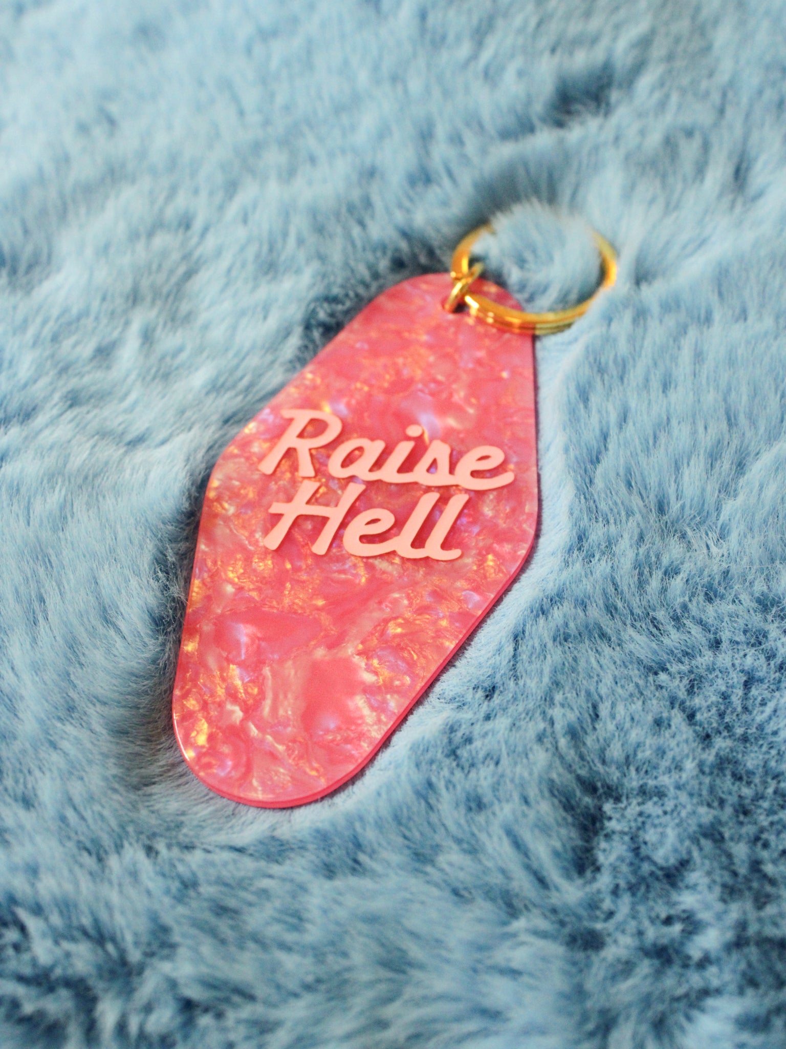 pink motel style keychain reads "raise hell" photographed on a fuzzy blue background